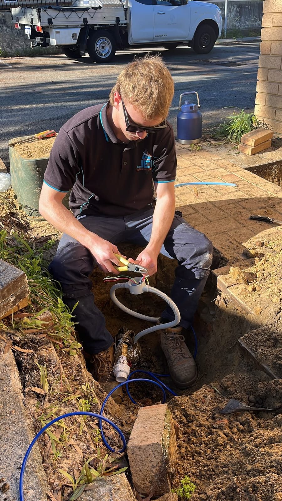 A JEC bore pump electrician in a branded Joondalup Electrical Contracting polo sitting at the edge of an open trench beside a paved driveway, stripping multi-core cable with yellow-handled strippers next to a junction box, grey flexible conduit and blue poly pipe.