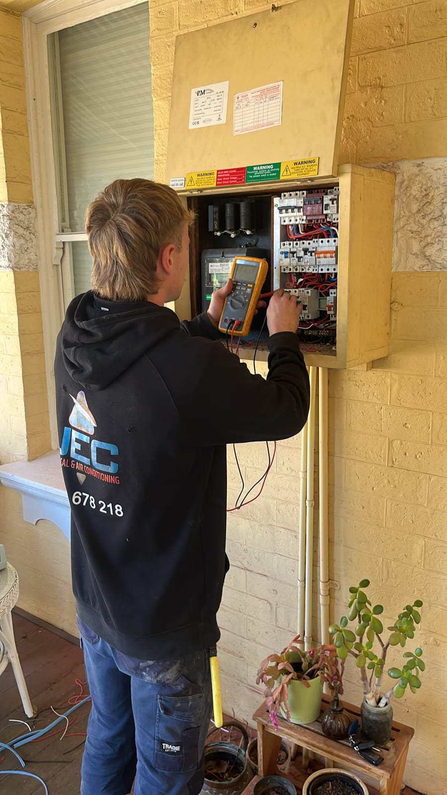 A JEC electrician in a branded JEC hoodie carrying out electrical fault finding at an open switchboard, testing the breakers with a yellow Fluke digital multimeter on red and black test leads at a limestone-brick house.