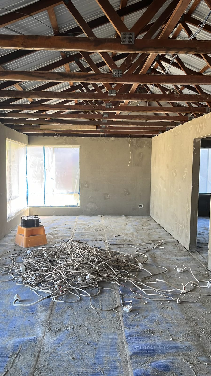 A house rewiring job in progress by JEC: a stripped-back room with bare plastered walls, exposed roof trusses and roof sheeting, and a pile of removed old cable and old switch mechanisms on the protected floor.
