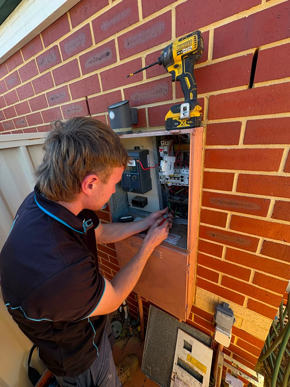 A JEC electrician part-way through a switchboard upgrade, working inside an open meter box on a red brick wall with a screwdriver in the board and an impact driver resting on top.