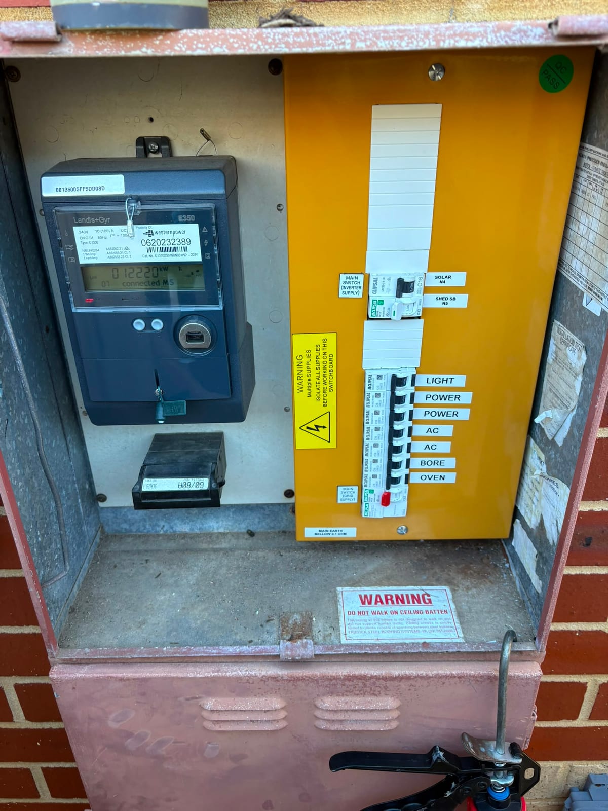 A finished switchboard upgrade by Joondalup Electrical Contracting: a Landis+Gyr smart meter beside a yellow-fronted board whose breakers are individually labelled LIGHT, POWER, AC, BORE and OVEN, with separate grid and inverter main switches and a green QC PASS sticker.