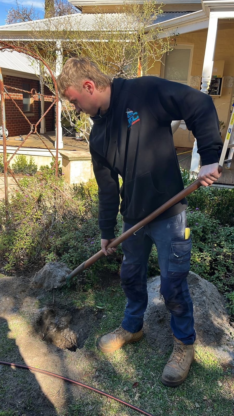 A JEC electrician in a branded Joondalup Electrical Contracting hoodie digging a cable trench with a long-handled shovel through a front lawn and garden bed, with orange conduit already run along the ground for a bore pump circuit.