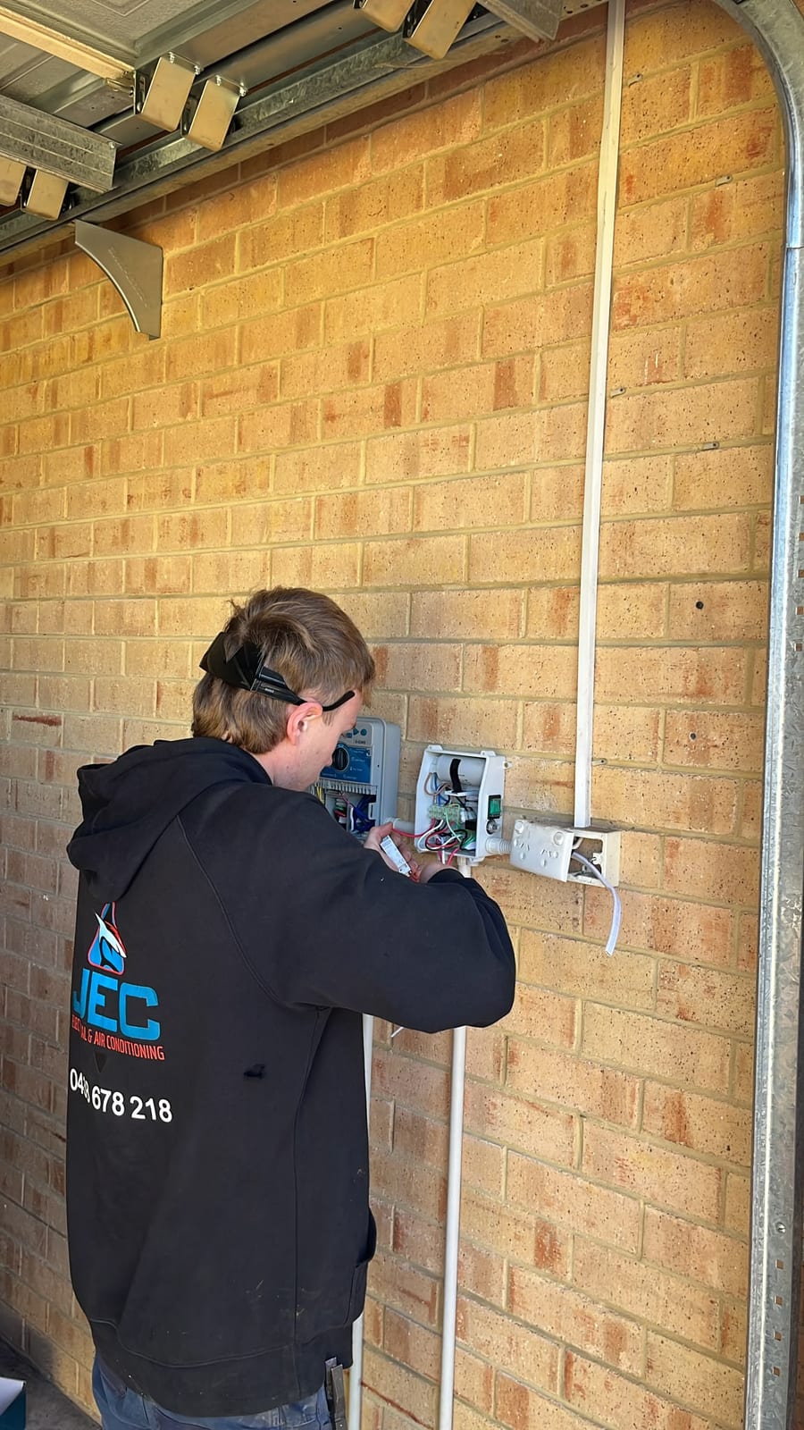 A JEC electrician in a branded Joondalup Electrical Contracting hoodie wiring an open weatherproof surface-mount outlet box during a power point installation, beside a wall-mounted irrigation controller on a brick garage wall with white surface conduit running up to it.